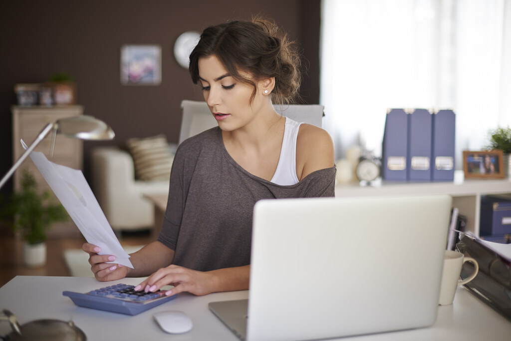 Como declarar imposto de renda: jovem de blusa cinza com o ombro de fora, segura uma folha com a mão direita enquanto digita em uma calculadora azul com a mão esquerda, está com o notebook aberto na sua frente e o fundo da imagem é sua sala, pois tem sofá almofadas e quadros em uma parede marrom